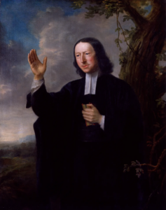 A picture of a John Wesley wearing a black robe and white collar holding a bible to his chest and holding his hand in the air as if preaching.